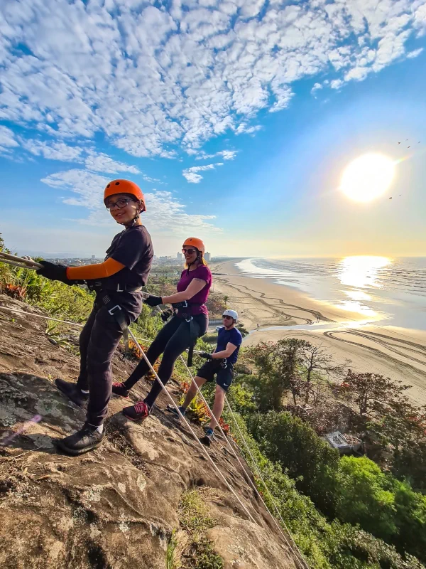 Família de turistas fazendo rapel em um paredão de pedra na Praia do Costão com o sol e o mar ao fundo.