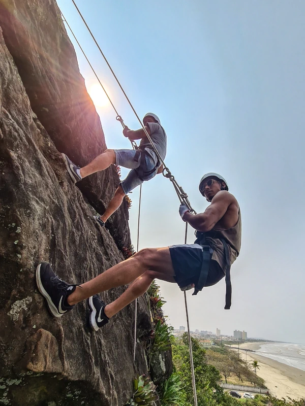 Grupo de turistas fazendo rapel em um paredão de pedra na Praia do Costão com o sol e o mar ao fundo.