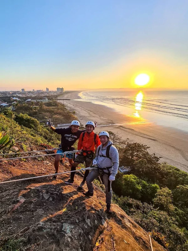 Grupo de turistas fazendo o Rapel do Nascer do Sol na Praia do Costão em Peruíbe com o sol nascendo ao fundo sobre o mar.